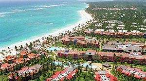 Aerial view of a tropical resort with red-roofed buildings, lush palm trees, and a stunning beach meeting turquoise ocean waters.
